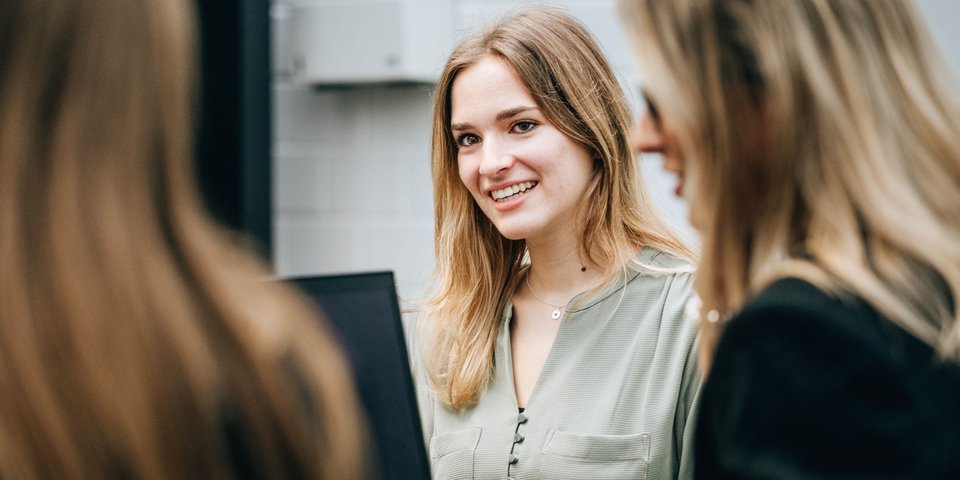 A young woman smiles and looks round as she sits with colleagues at a table with a laptop.