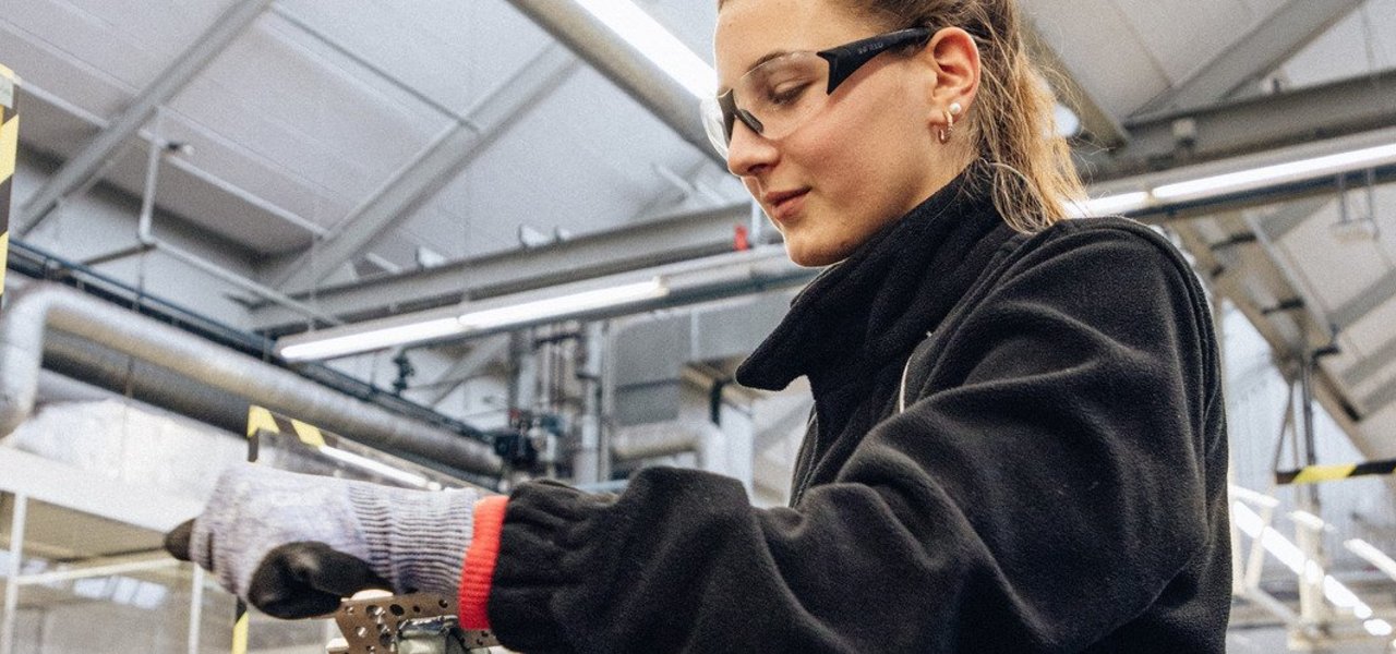 A technical apprentice wearing safety goggles and safety gloves works on a vice in a workshop environment.