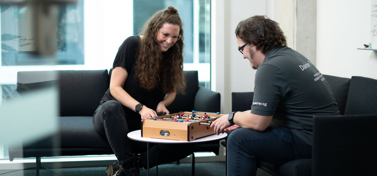 Two employees sit on a sofa in the Daimler Truck Campus and play mini table football together.