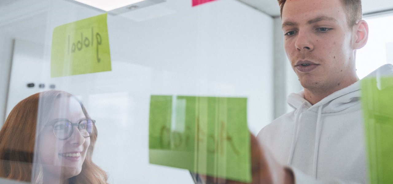 Two young employees stand behind a glass wall with colourful sticky notes, the woman smiles while the man arranges a note.