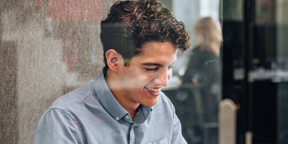 A young commercial trainee smiles as he works on his laptop in a glazed meeting room.
